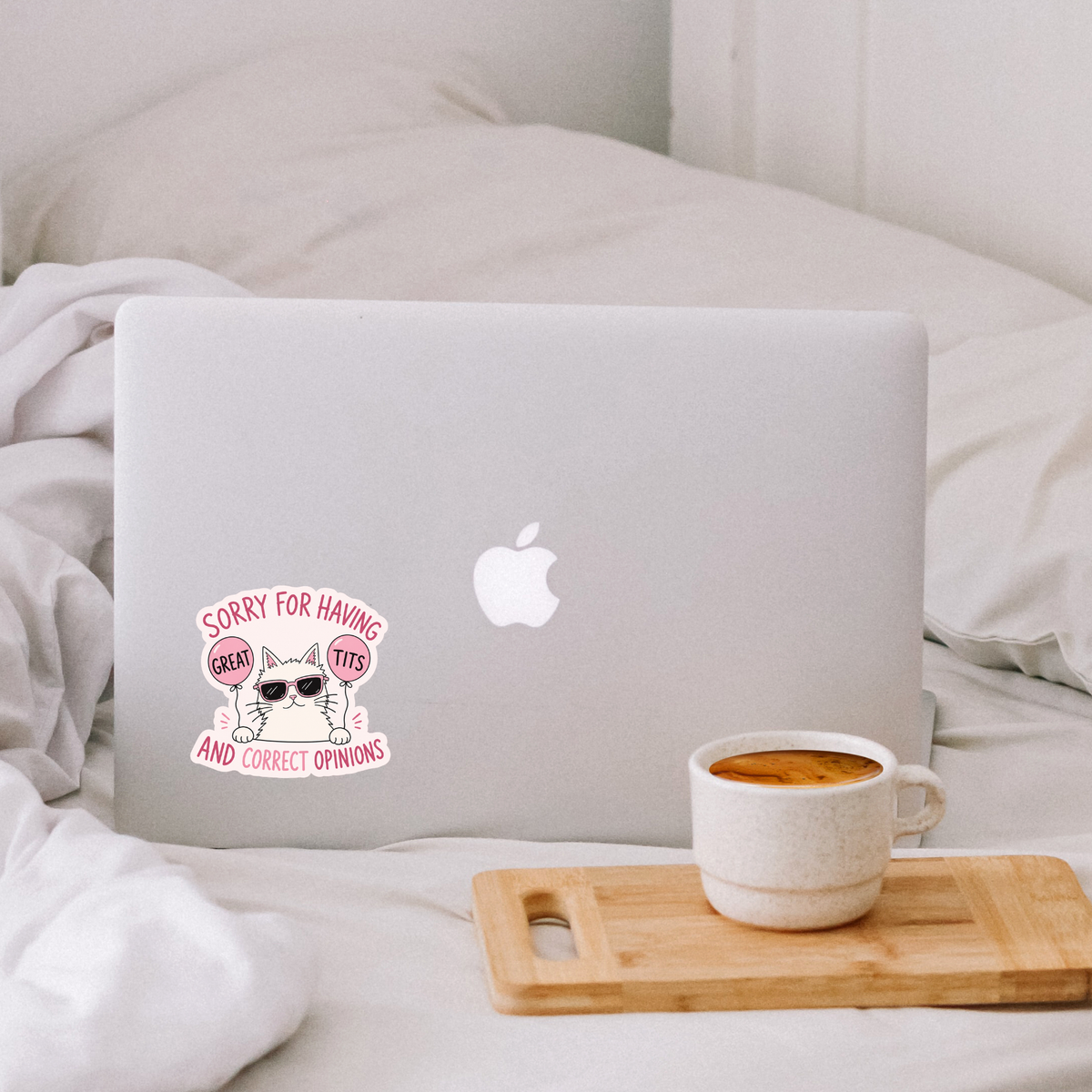 A white sticker with a cartoon cat wearing sunglasses and the text 'SORRY FOR HAVING GREAT FITS AND CORRECT OPINIONS' applied to the back of a silver laptop on a bed. A wooden tray holding a cup of coffee sits in the foreground.
