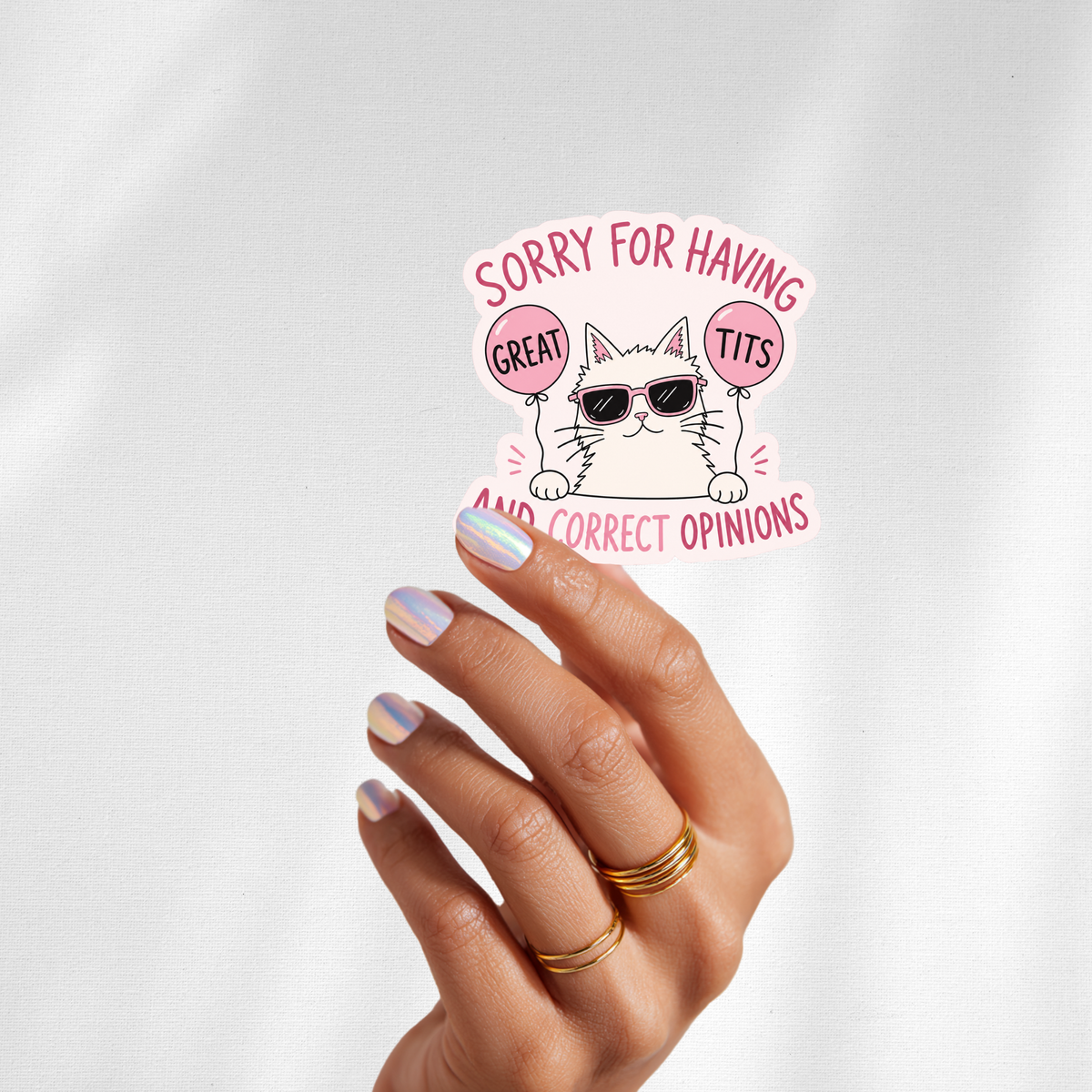 A hand with iridescent nail polish and gold rings holds a die-cut sticker against a white fabric background. The sticker features a cartoon white cat wearing sunglasses and holding pink balloons that read 'GREAT TITS', with the text 'SORRY FOR HAVING' above and 'AND CORRECT OPINIONS' below.