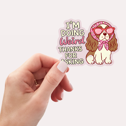 Sticker with a dog wearing sunglasses and a bandana, text 'I'm doing weird thanks for asking', held by a hand.