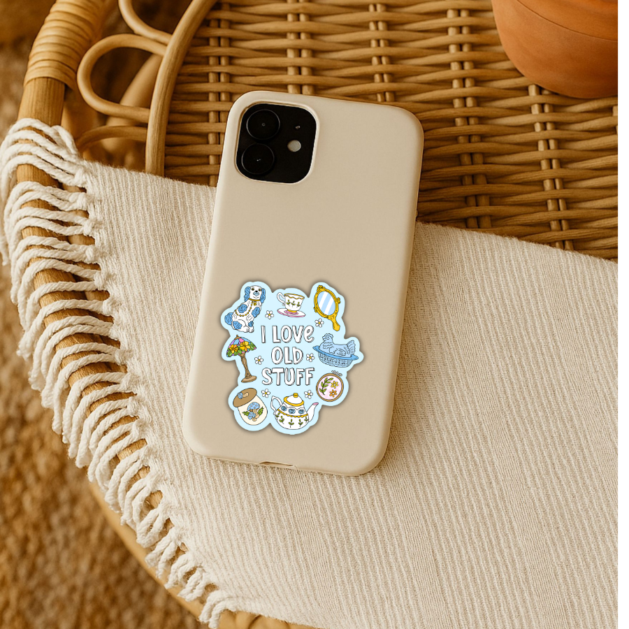 A beige phone case with a colorful retro-style sticker that reads 'LOVE OLD STUFF' with cartoon illustrations, resting on a cream fringed textile inside a wicker basket.