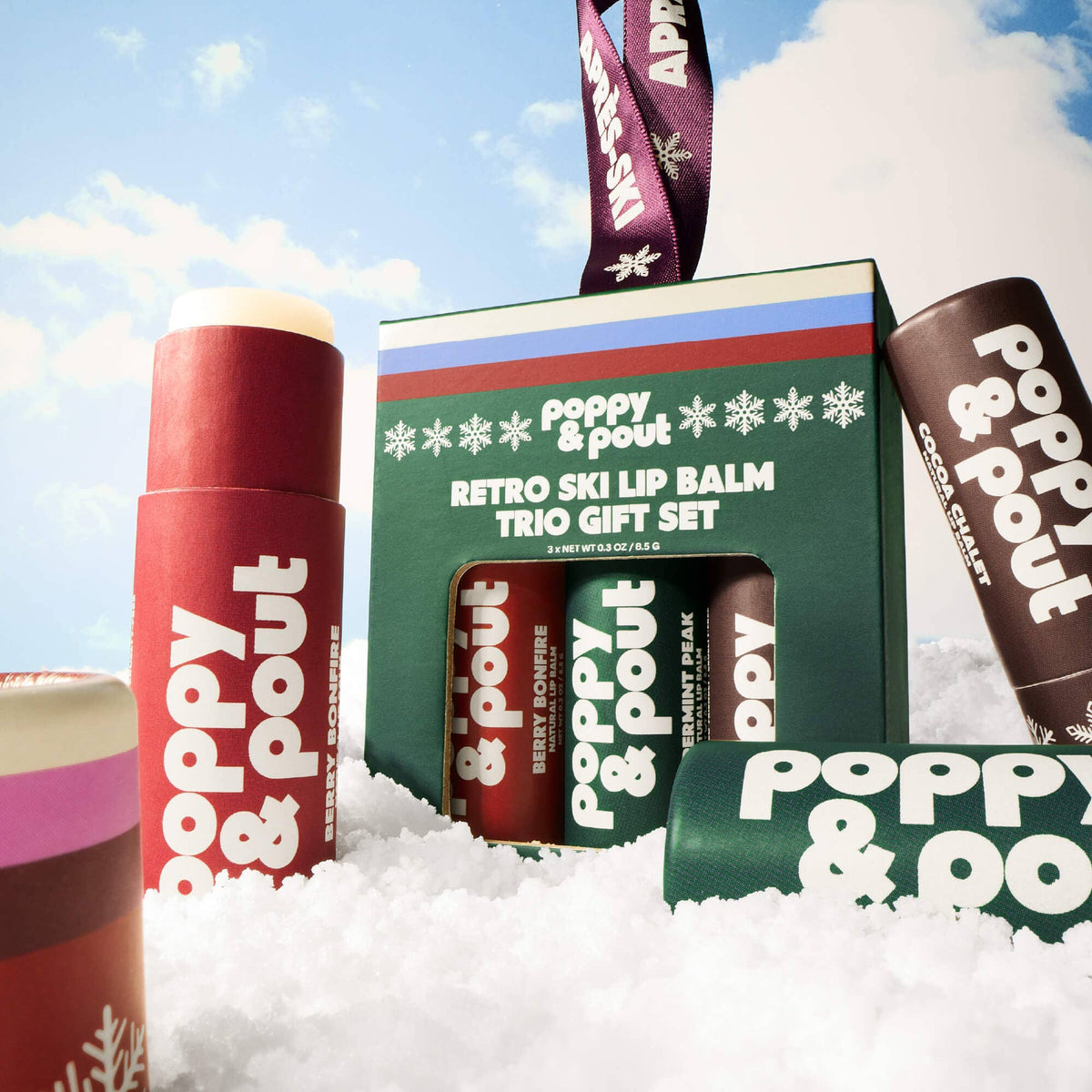 Poppy & Pout Retro Ski Lip Balm Trio Gift Set displayed in a snowy setting under a blue sky. The set includes a green gift box with a hanging ribbon and three lip balms in red, brown, and pink packaging with white lettering.